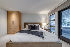 A modern bedroom with a large bed, wooden wardrobe, bedside table with lamp, and a view of a snowy landscape through large windows. - This is the largest ski in ski out rental townhome in Whistler. And it's swanky. (Whistler)