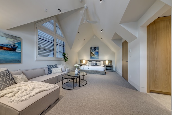 A modern bedroom with a loft-style ceiling, a large window with blinds, a bed with a nightstand, and a seating area with a sofa and coffee table.