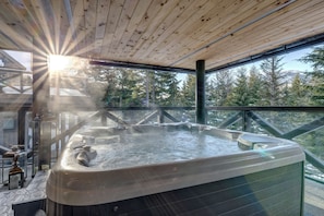 A hot tub with steam rising, surrounded by a wooden deck and glass railing, overlooking a forested area. - This is the largest ski in ski out rental townhome in Whistler. And it's swanky. (Whistler)