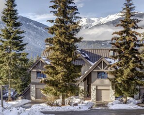 A two-story house with a stone facade, large windows, and a garage. It is surrounded by snow-covered ground and trees, with mountains in the background. - Pinnacle Ridge 3 - Ski in Ski out, 7 Bedrooms, Hot Tub, Sauna & Billiards (Whistler)