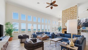 A spacious living room with a large window overlooking the ocean, a ceiling fan, and a variety of seating arrangements. - Manu Kai - Beachfront House With Private Pool And Hot Tub 6 Bedroom Home by RedAwning (South Padre Island)