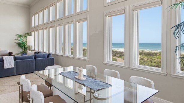A spacious room with a large glass table, white chairs, a blue sofa, and a view of the beach through large windows. - Manu Kai - Beachfront House With Private Pool And Hot Tub 6 Bedroom Home by RedAwning (South Padre Island)