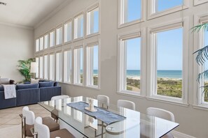 A spacious room with a large glass table, white chairs, a blue sofa, and a view of the beach through large windows.