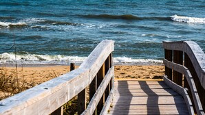 Ein hölzerner Steg führt zu einem Sandstrand, im Hintergrund das Meer.