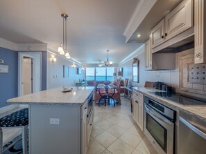 A modern kitchen with a central island, built-in appliances, and a dining area with a view. - The Grande Pelican | Oceanfront + Community Pool! (Carolina Beach)