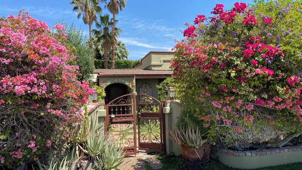 A garden entrance with a wrought iron gate, a pathway leading to a building with a decorative archway, and various flowering plants. - Palm Springs Paradise (Palm Springs)