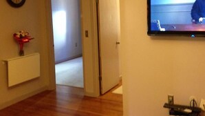 Un passadís amb sòl de fusta, un rellotge a la paret, un televisor penjat a la paret i una porta que dona a una altra habitació.
