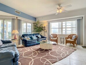 Ein Wohnzimmer mit einem blauen Sofa, einem Glastisch, einem gemusterten Teppich und zwei Korbstühlen.