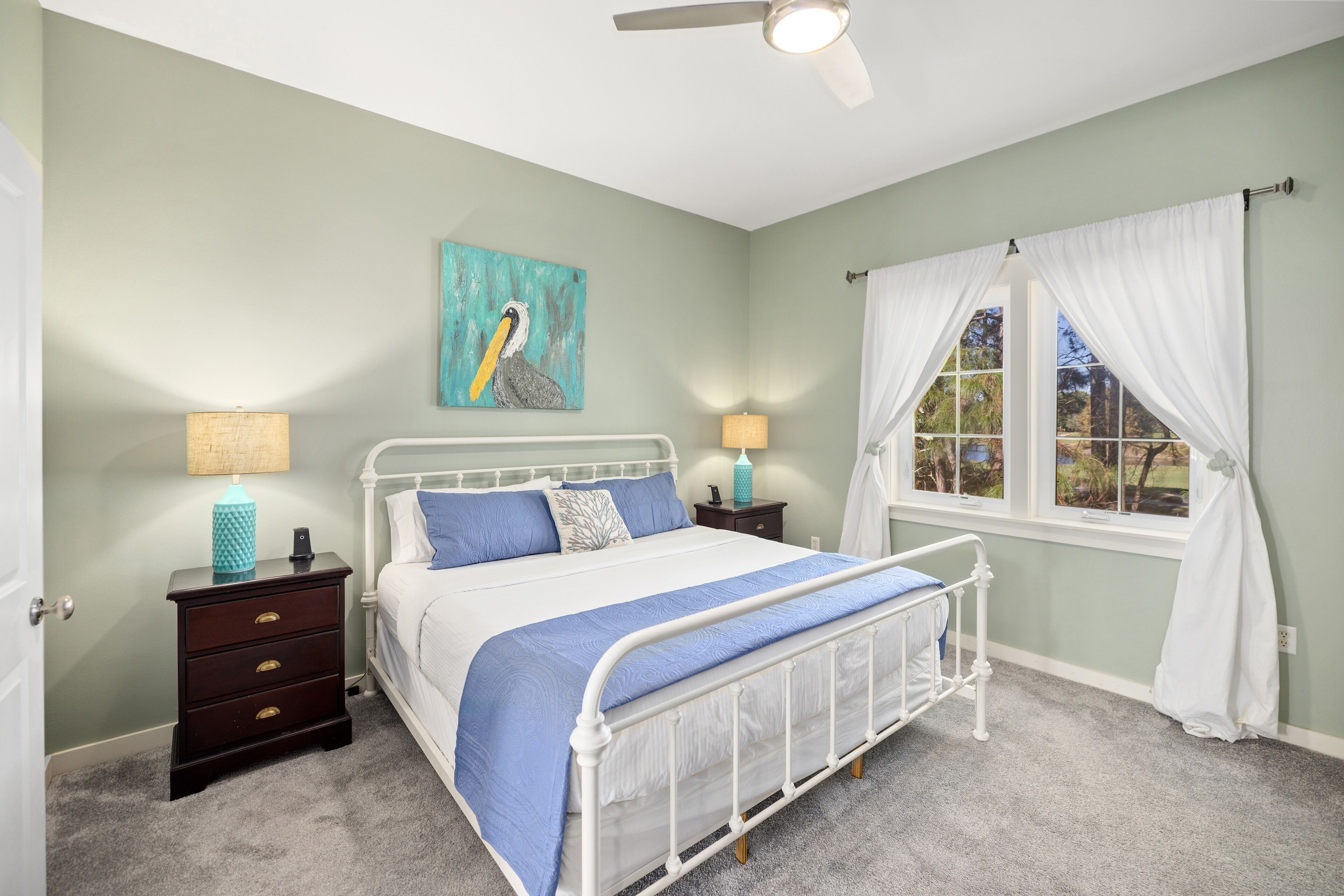 Un dormitorio con estructura de cama de metal blanco, colcha azul, dos mesitas de noche con lámparas, un cuadro de pared color turquesa y una ventana con cortinas blancas.