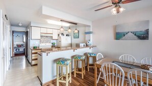 A modern kitchen with a central island, wooden flooring, and a dining area with white chairs. - Sea Colony 2A | Oceanfront, Private Beach Walkway (Carolina Beach)