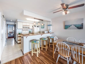 A modern kitchen with a central island, wooden flooring, and a dining area with white chairs. - Sea Colony 2A | Oceanfront, Private Beach Walkway (Carolina Beach)