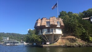 Une structure unique, ressemblant à une maison et surmontée d’un drapeau, située au bord d’un plan d’eau.