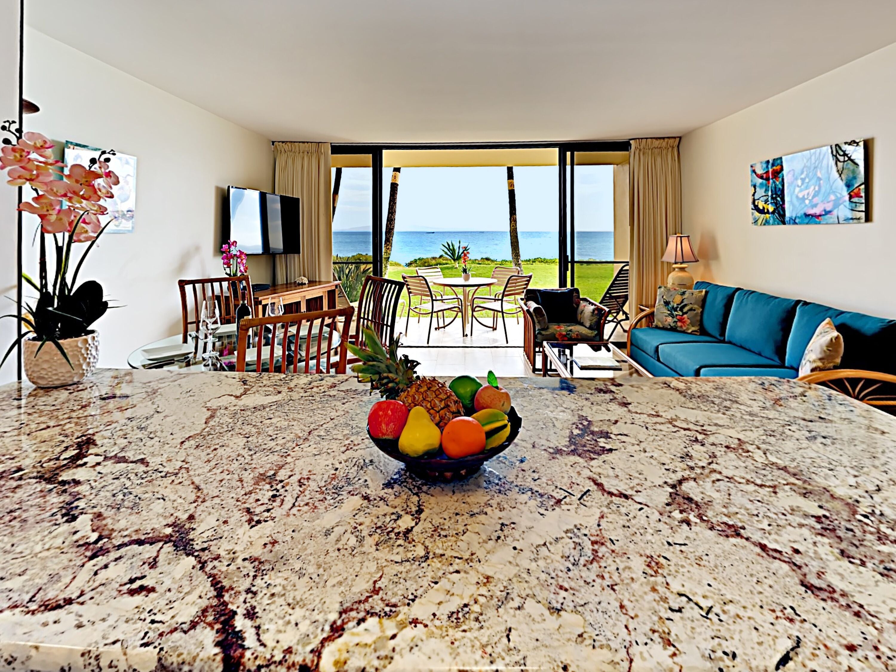 A modern hotel room with a marble dining table, a fruit bowl, a blue sofa, and a balcony with a view of the ocean.