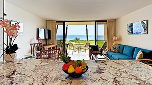Una habitación de hotel moderna con una mesa de comedor de mármol, un frutero, un sofá azul y un balcón con vistas al océano.
