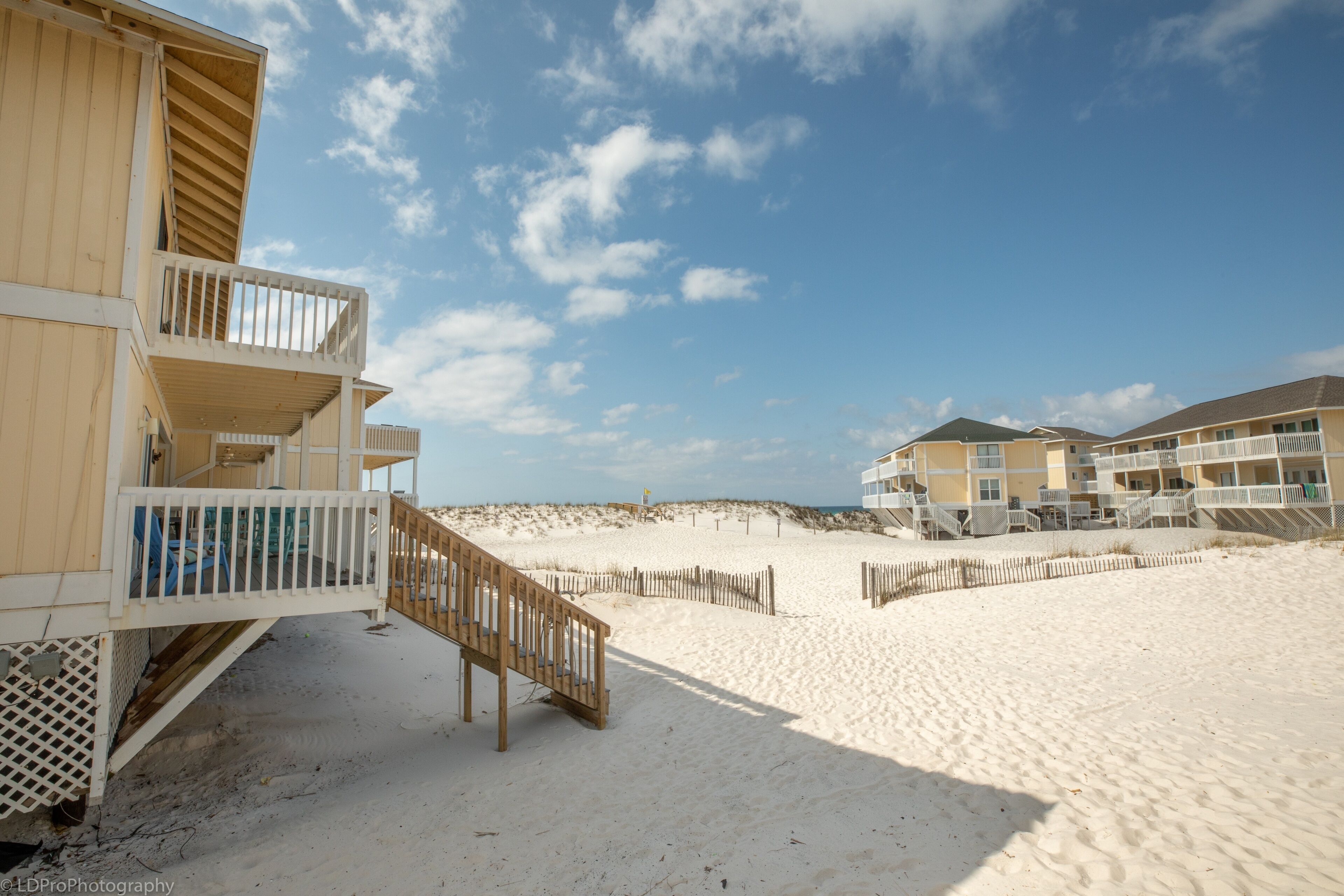 A beachfront hotel with a wooden staircase leading to a balcony, sandy beach, and a clear sky.