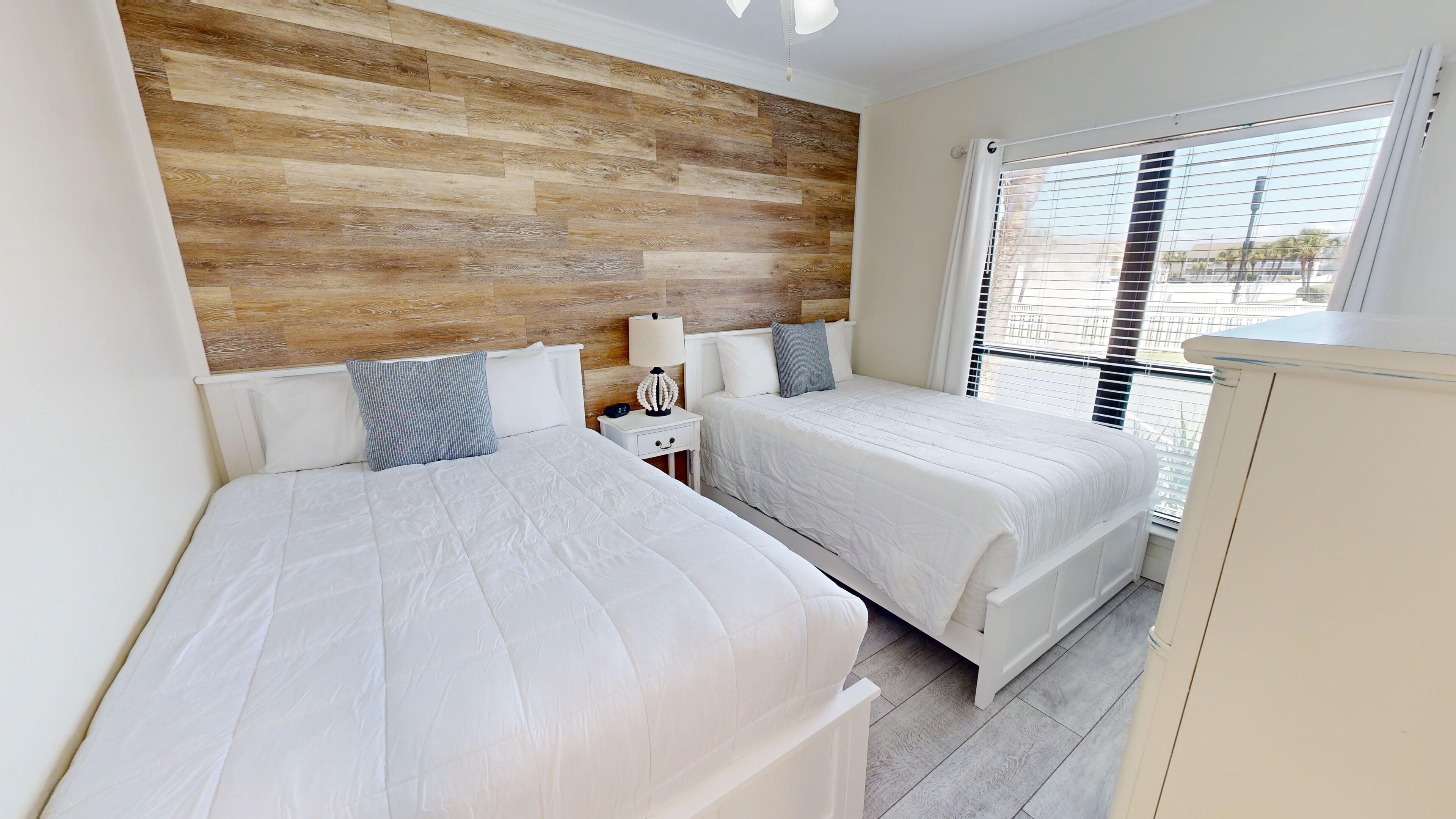 A bedroom with two beds, a wooden headboard, a nightstand with a lamp, and a window with blinds.