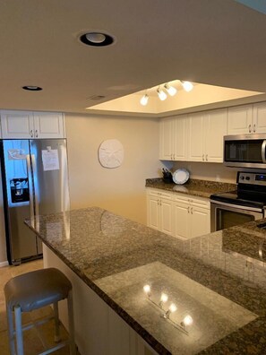 Cozinha moderna com armários brancos, bancadas de granito e eletrodomésticos de aço inoxidável, além de uma ilha central com banquetas.