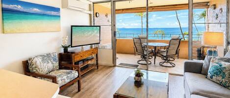 A living room with a TV, wooden furniture, a glass table, and a balcony with a view of the ocean.