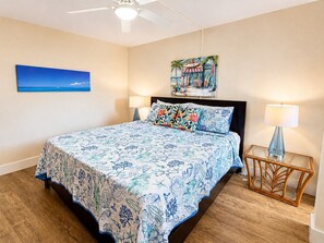 Une chambre à coucher avec un lit, deux tables de chevet, un ventilateur de plafond, un tableau et une vue sur l’océan.
