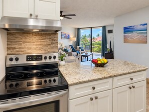 A modern kitchen with a stainless steel range, a granite countertop, and a dining area with a view of the outdoors. - Scenic Kihei Village by the Sea Renovated Condo Ocean Views Lanai Pool (Kihei)
