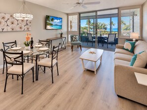 A spacious living area with a dining table, chairs, a sofa, and a view of the ocean through large windows. - Scenic Kihei Village by the Sea Renovated Condo Ocean Views Lanai Pool (Kihei)