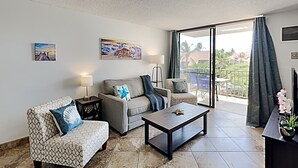 En stue med sofa, lænestole, sofabord og et tv-bord. Der er to billeder på væggen og en balkon med udsigt.