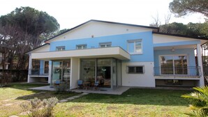 A two-story house with a flat roof, a balcony, and a covered patio area with outdoor seating. - Coastal villa in Lignano Riviera (Lignano Sabbiadoro)