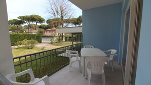 A balcony with white plastic chairs and a table, overlooking a garden with trees and a pathway. - Coastal villa in Lignano Riviera (Lignano Sabbiadoro)