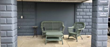 A covered outdoor seating area with two wicker chairs and a small stool, under a building with a grey brick wall and a 'PARKING' sign indicating high and low tide directions.