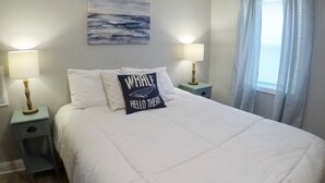 A neatly made bed with white bedding and two bedside lamps. A pillow with the text "WHALE HELLO THERE" is placed on the bed. - Sea Level Sanctuary's Low Tide in Seaside Park (Seaside Park)