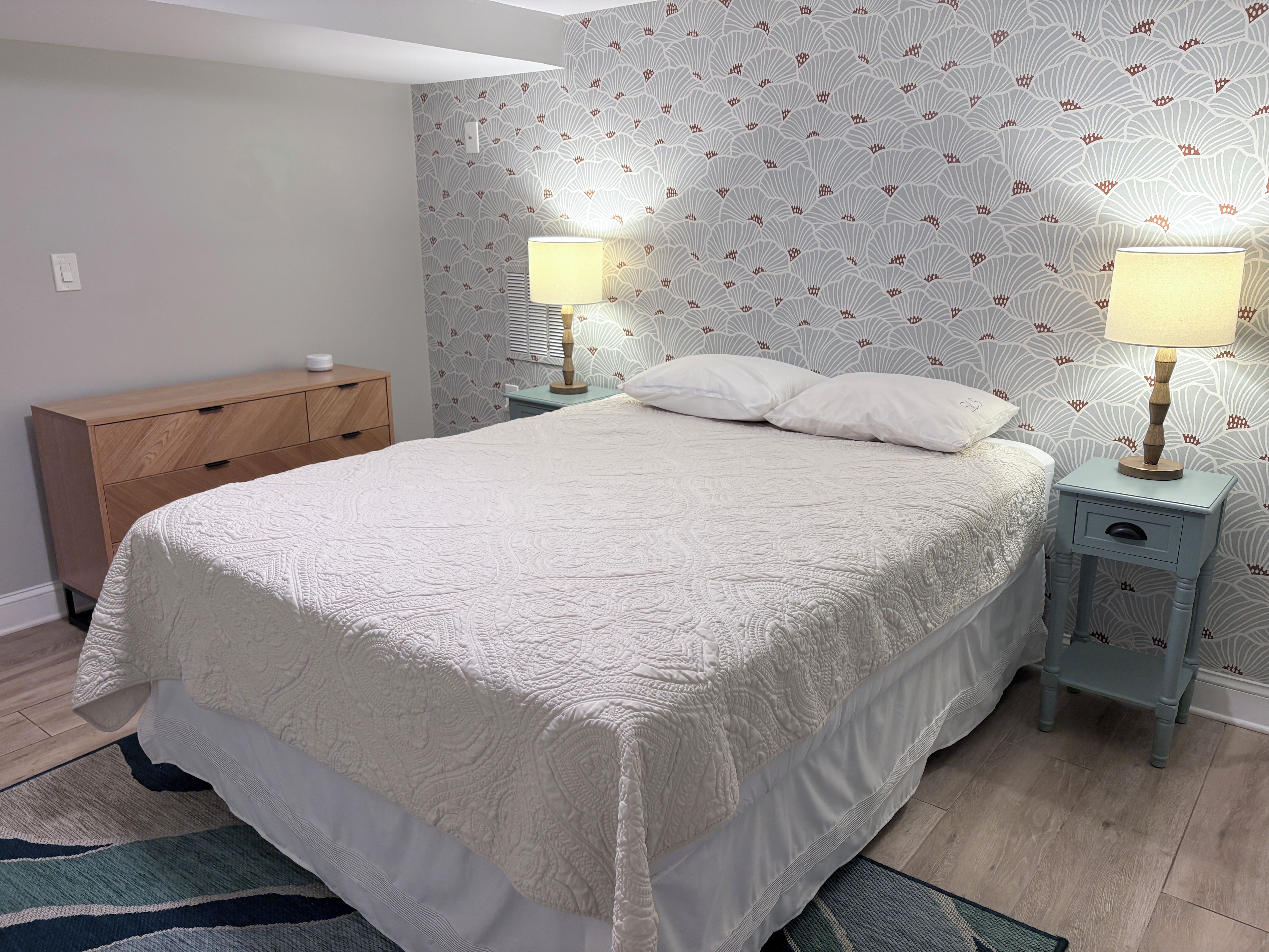 A bedroom with a large bed, two bedside tables with lamps, a wooden dresser, and patterned wallpaper.
