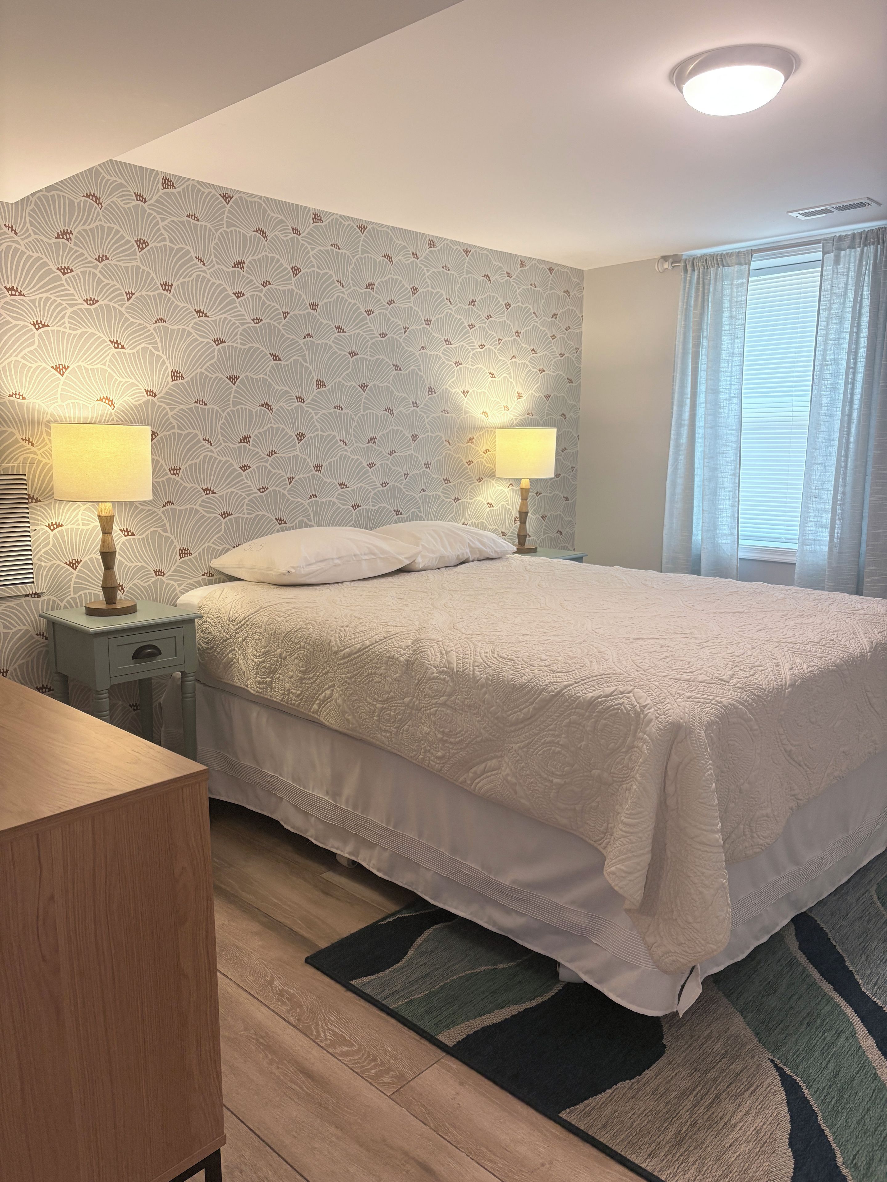 A hotel room with a bed, two bedside lamps, a nightstand, a wooden dresser, and a window with curtains.