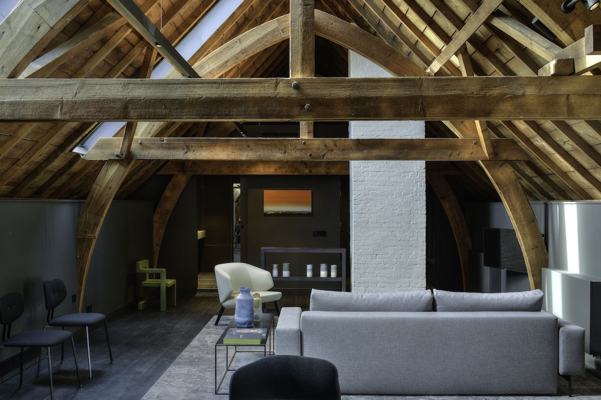 An attic room with exposed wooden beams, a grey sofa, a white armchair, and a small table with a vase.
