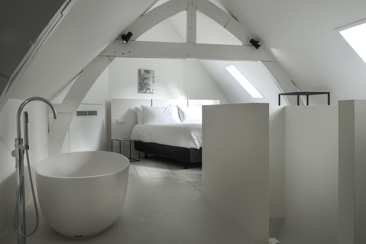Quarto sótão moderno, com decoração em tons de branco, claraboia, banheira grande e cama com roupa de cama branca.