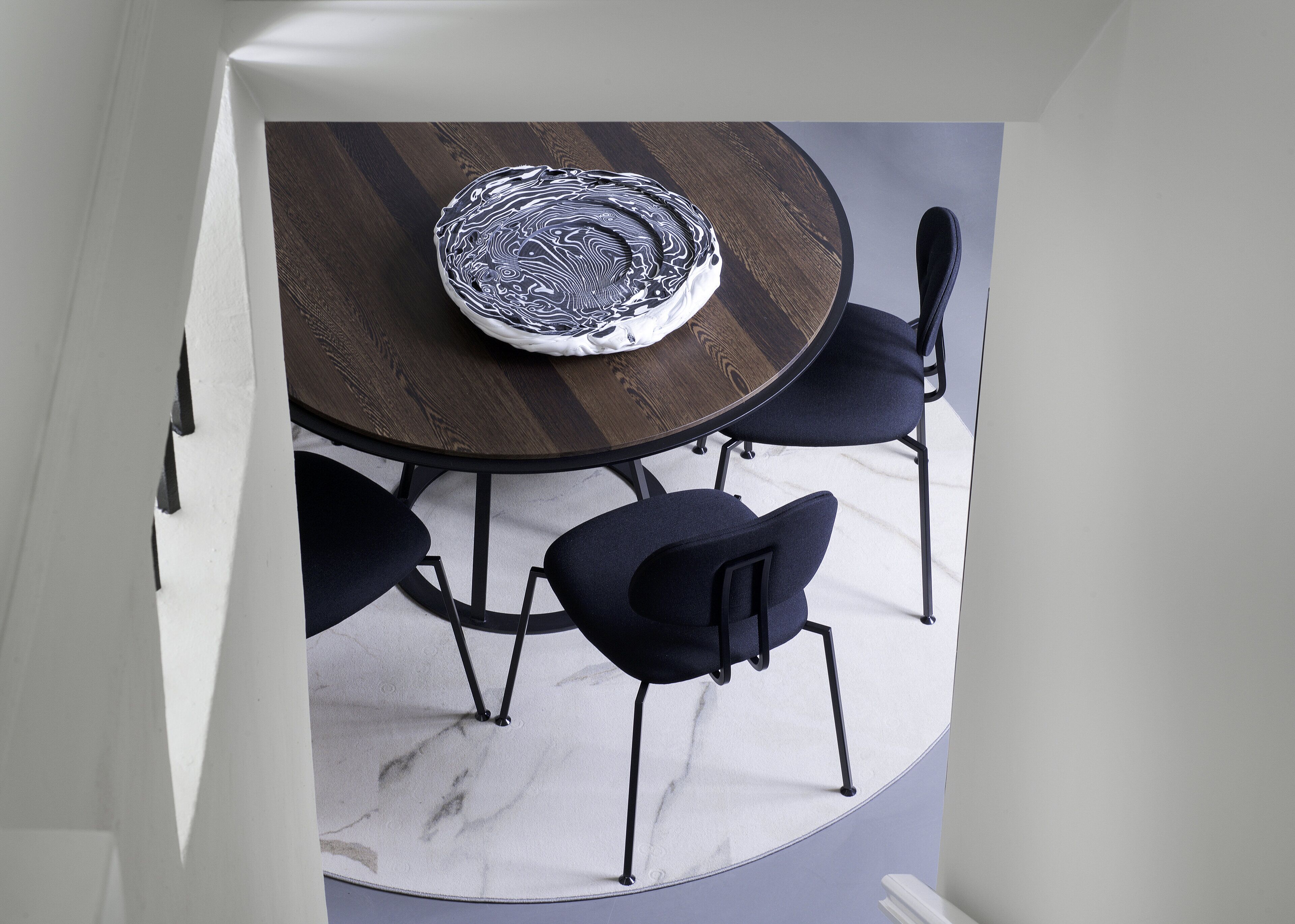 Una mesa redonda de comedor con acabado en madera oscura y un centro de mesa blanco y texturizado. Cuatro sillas azul oscuro con patas metálicas la rodean.