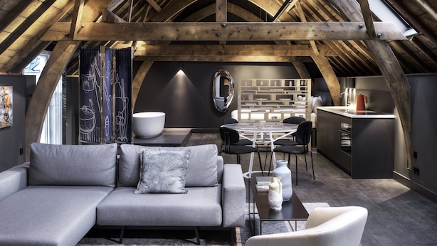 An attic-style room with a wooden ceiling, a gray sofa, a dining table with chairs, and a kitchen area. - Hotel Kazerne (Eindhoven)