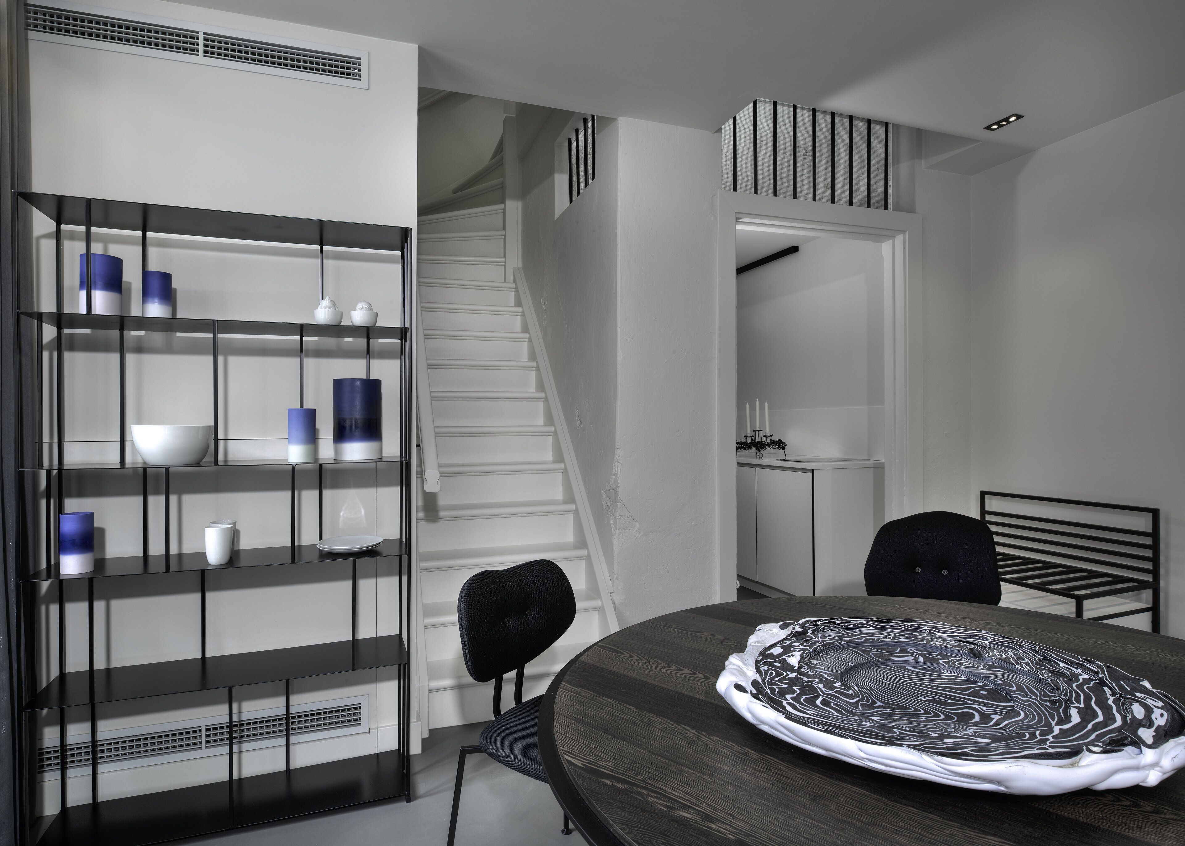 A modern dining area with a round table, black chairs, and a decorative plate. A staircase leads to a second floor, and a black metal shelf with decorative items is visible.