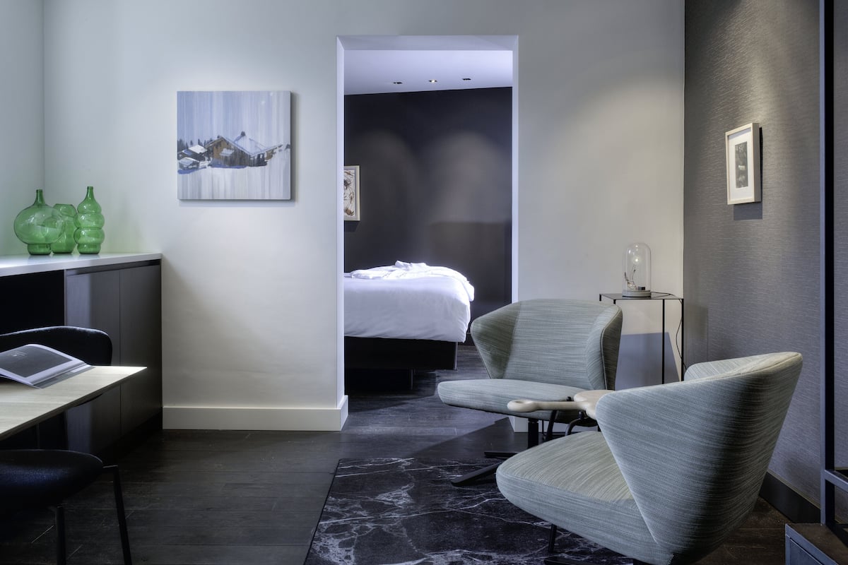 A modern hotel room with a bed, a desk, a chair, and a small table. There is a painting on the wall and a vase on the desk.