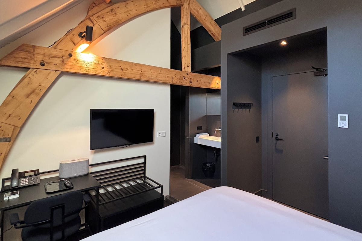 A modern hotel room with a wooden ceiling, a flat-screen TV, a desk with a chair, a bed, and a bathroom with a sink and toilet.