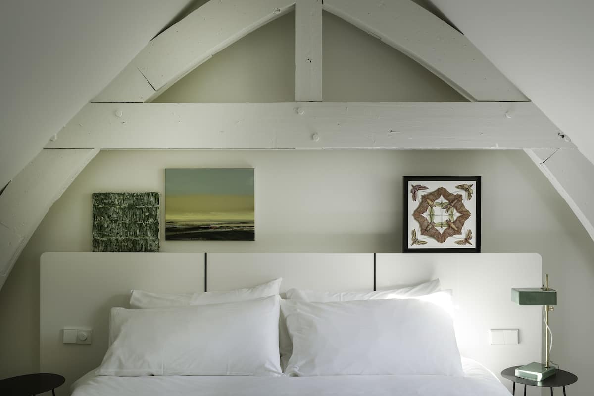 A bedroom with a bed, two pillows, a nightstand with a lamp, and two wall-mounted artworks.