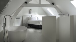 A modern, white-themed attic bedroom with a skylight, a large bathtub, and a bed with white linens. - Hotel Kazerne (Eindhoven)