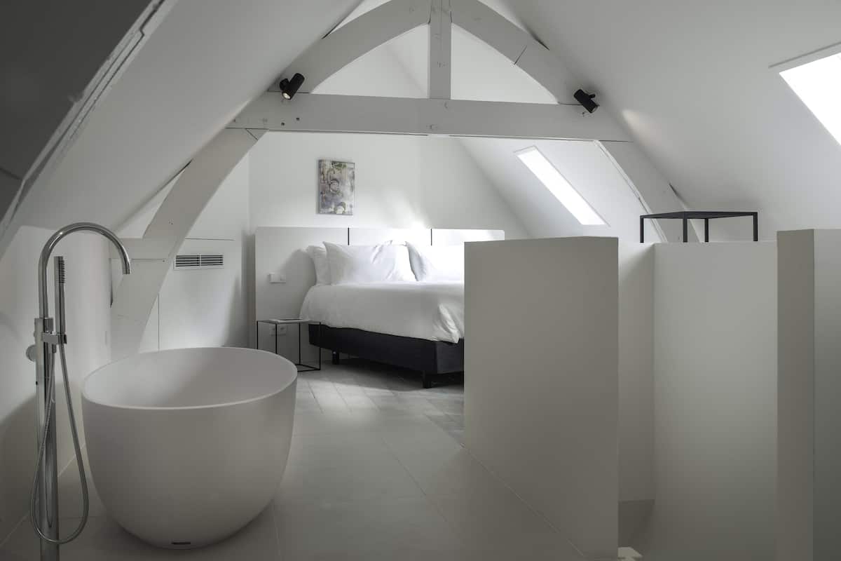 A modern, white-themed attic bedroom with a skylight, a large bathtub, and a bed with white linens.
