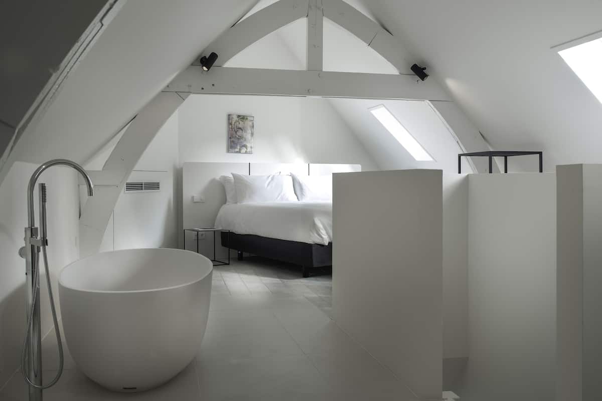 A modern, white-themed attic bedroom with a skylight, a large bathtub, and a bed with white bedding.