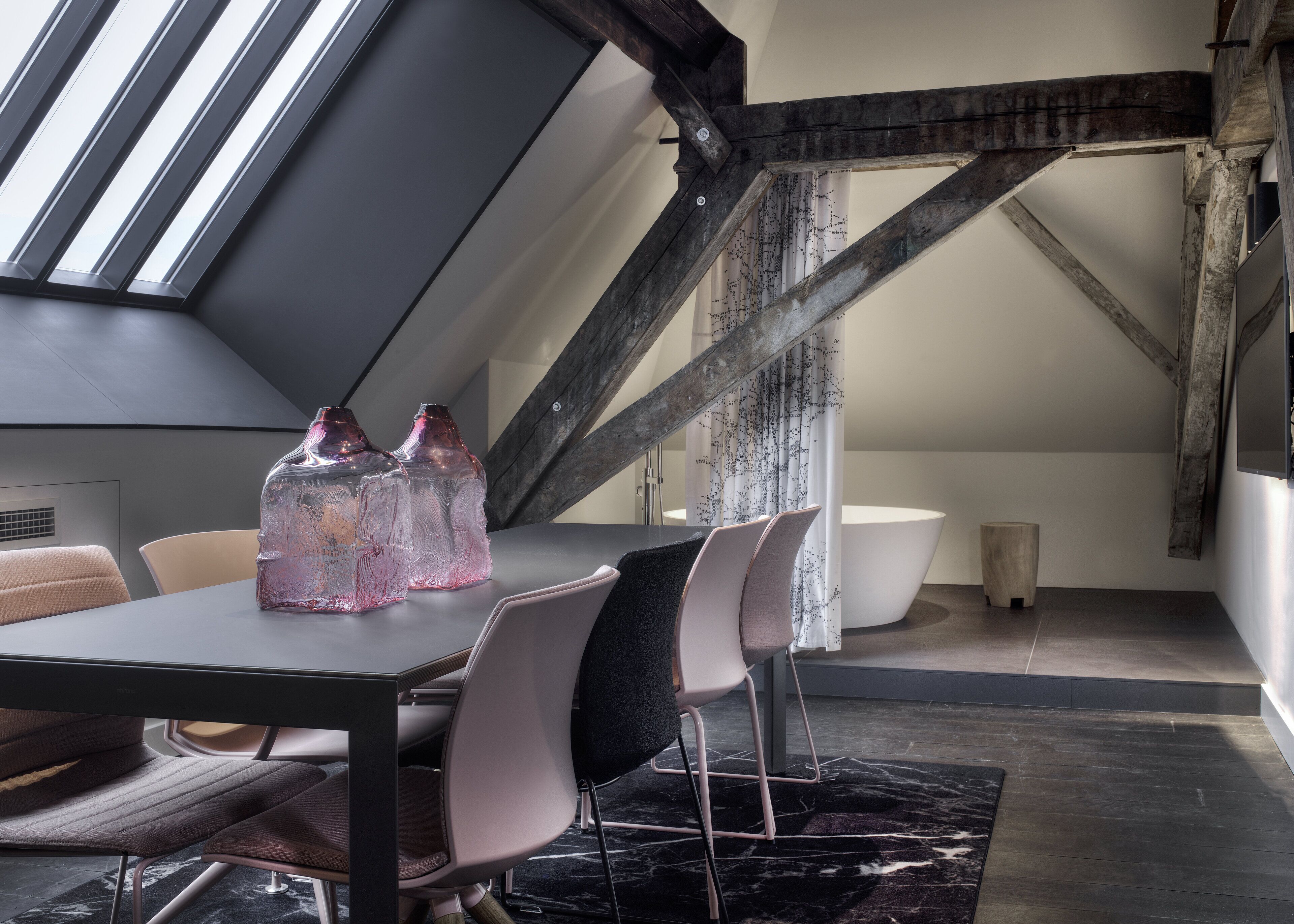 Ein modernes Schlafzimmer im Loft-Stil mit schrägverdeckter Decke, freiliegenden Holzbalken, einem Oberlicht, einem großen Fenster, einer runden Badewanne und einem Essbereich mit Stühlen und Tisch.
