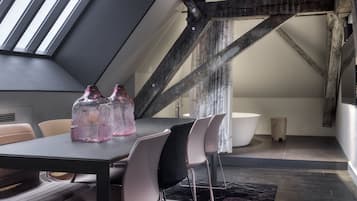 Ein modernes Schlafzimmer im Loft-Stil mit schrägverdeckter Decke, freiliegenden Holzbalken, einem Oberlicht, einem großen Fenster, einer runden Badewanne und einem Essbereich mit Stühlen und Tisch.