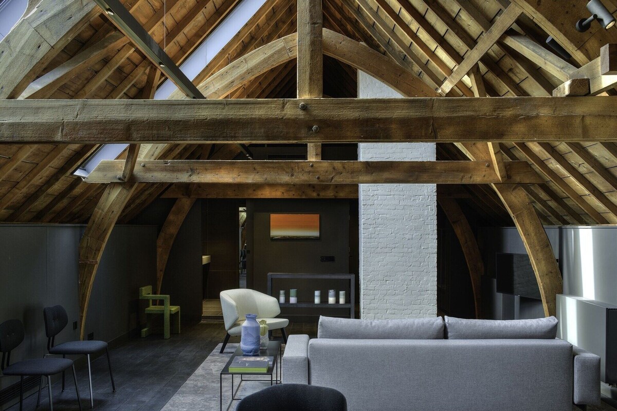 An attic room with exposed wooden beams, a grey sofa, a white chair, and a small table with a vase.
