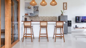 A modern kitchen with three wicker pendant lights, a built-in bar with three wooden barstools, a refrigerator, and a flat-screen TV mounted on the wall.