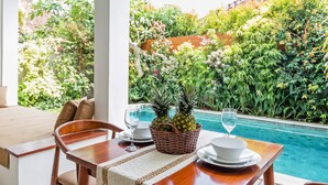 A dining table with a wicker basket containing two pineapples, white plates, and wine glasses, set against a backdrop of a lush garden and a swimming pool.