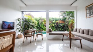 A modern living room with a sofa, a coffee table, and a dining area with chairs and a table. There is a TV on the wall and a large window with a view of greenery outside.