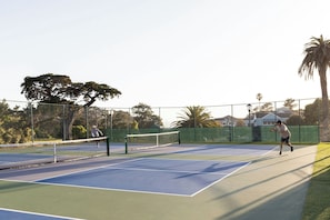 Una cancha de tenis al aire libre con red, rodeada por una valla, árboles y edificios al fondo.